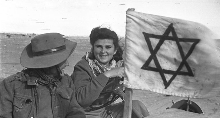 Two smiling Israeli fighters seated beside a hand-painted Star of David flag in the desert during the 1948 war.