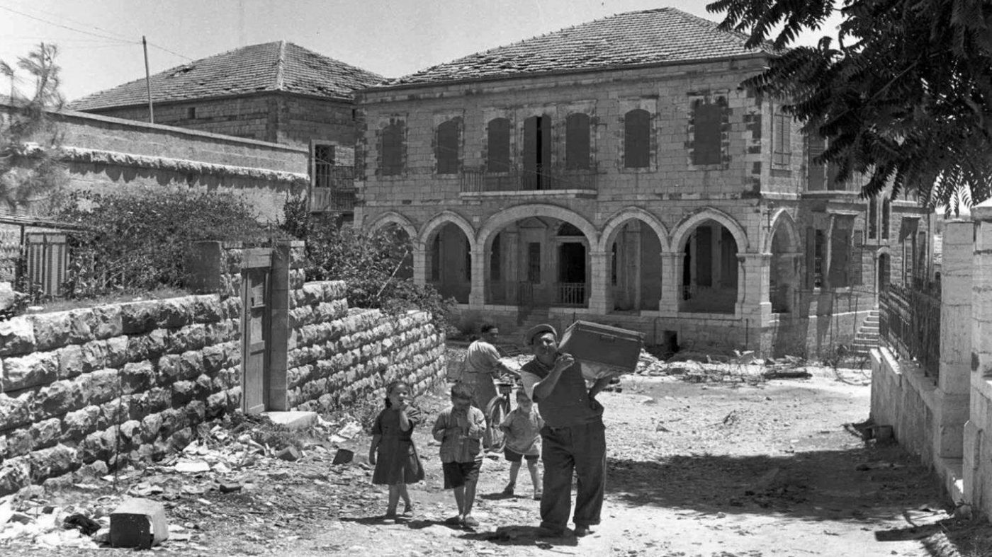 A family carries belongings past damaged stone buildings in a Jewish neighborhood during the 1948 war.